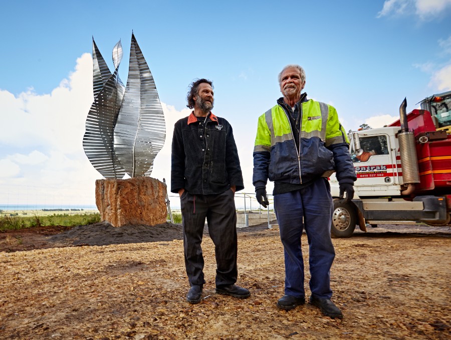 Two men stand in front of a large silver sculpture