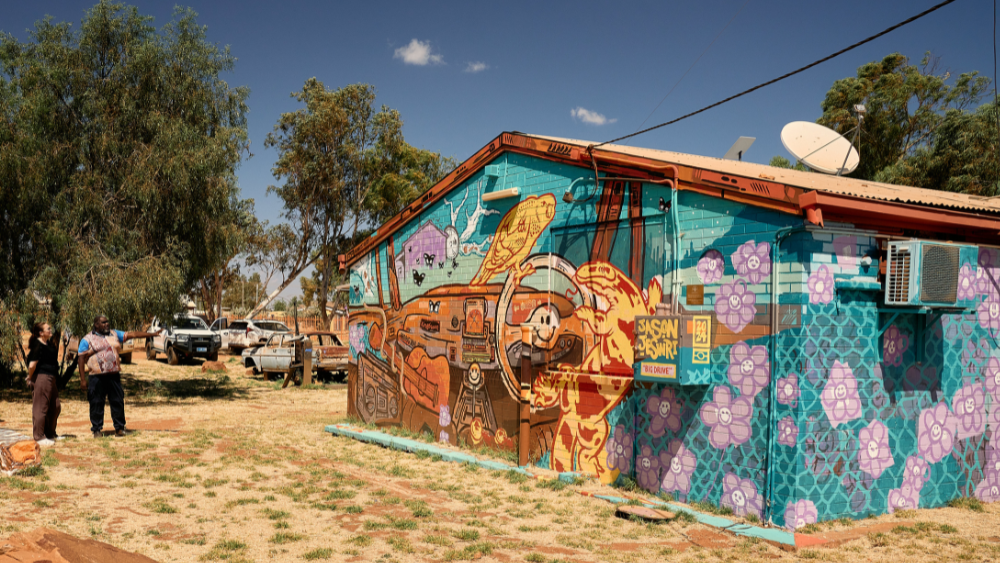 Two people looking at a building in an outback town, painted with a mural of a car being driven by a lizard and a bird.