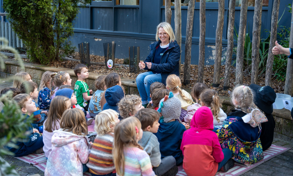 A teacher, smiling, sitting outside, speaking to a group of primary school students.